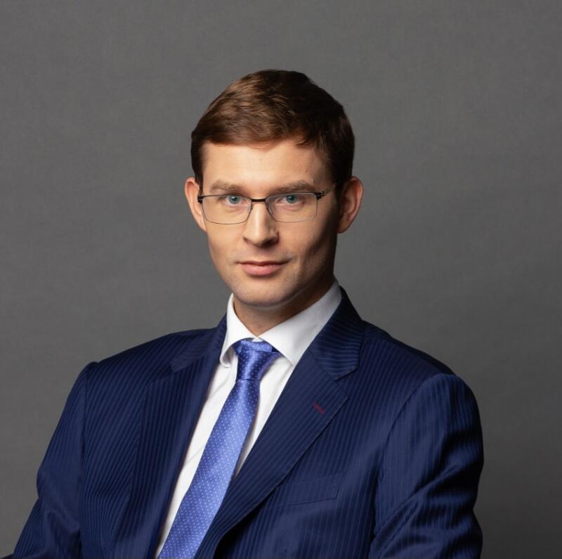 A man with glasses is wearing a blue suit and tie against a gray background.
