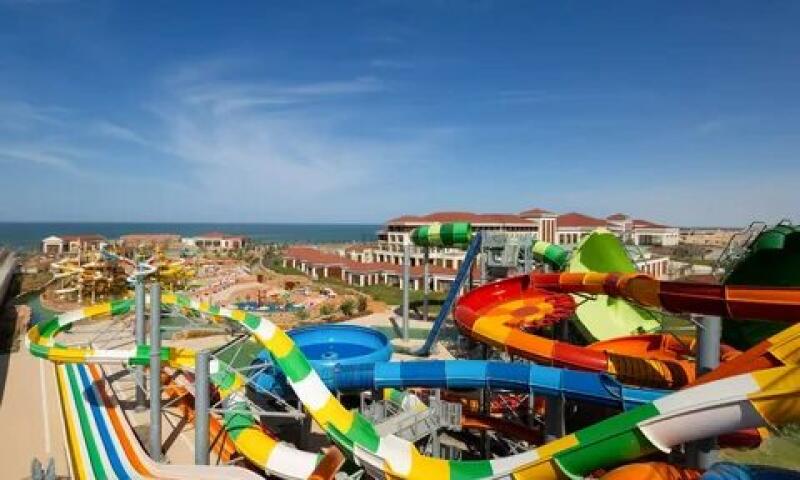 An aerial view shows a water park featuring various colorful slides, buildings, and the ocean under a clear blue sky.