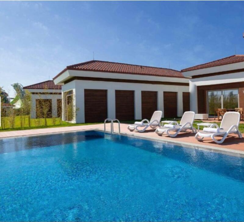 The image shows a swimming pool in front of a building with lounge chairs. The building has a white exterior with brown accents and a red tile roof.