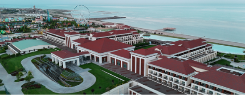 An aerial view of a large hotel complex with red roofs and a nearby amusement park featuring a Ferris wheel, all set against a backdrop of the sea.