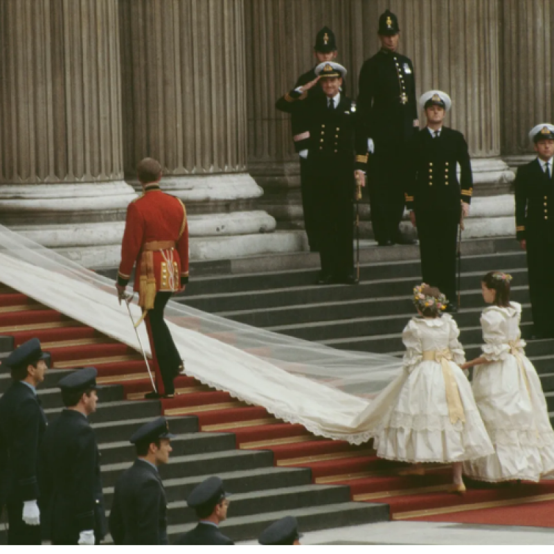 A man in a red military uniform walks up the stairs, followed by two young girls in white dresses carrying a long white veil; servicemen in navy uniforms stand at the top of the stairs.