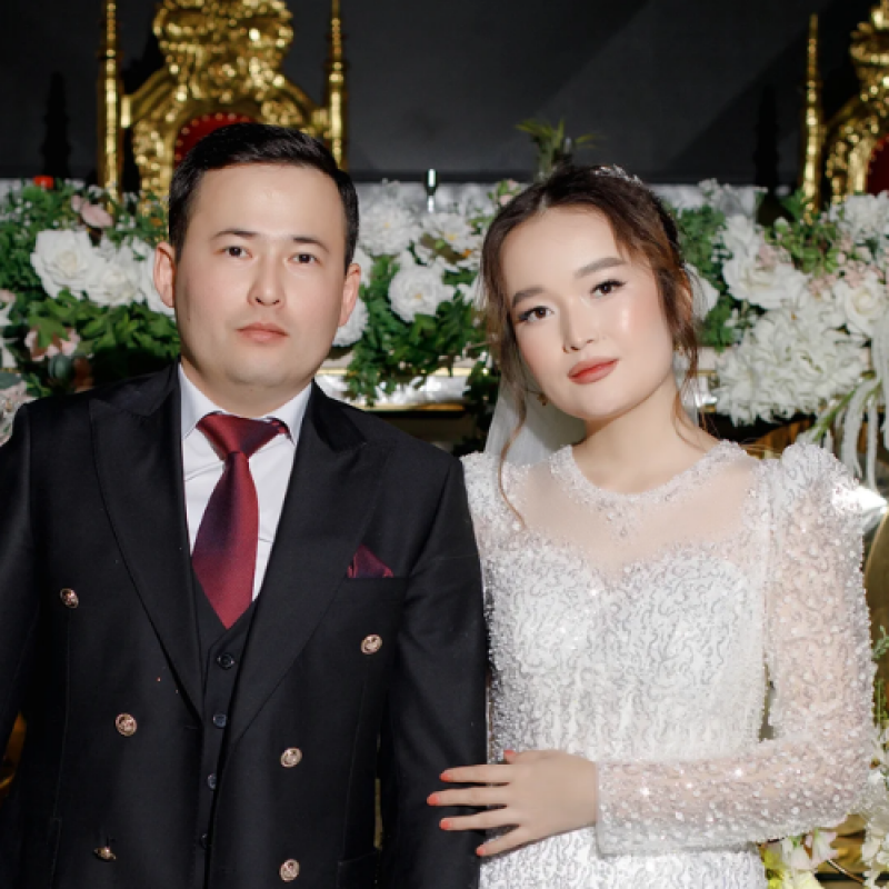 A wedding photo shows a groom in a black suit with a red tie and a bride in a white dress with long sleeves, posing in front of a floral arrangement and a golden decorative structure.