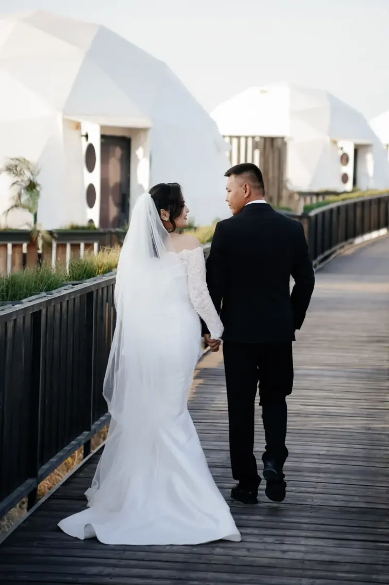 A bride in a white off-the-shoulder dress and veil walks hand-in-hand with a groom in a black suit on a wooden walkway, looking back at him. White dome-shaped buildings are visible in the background.
