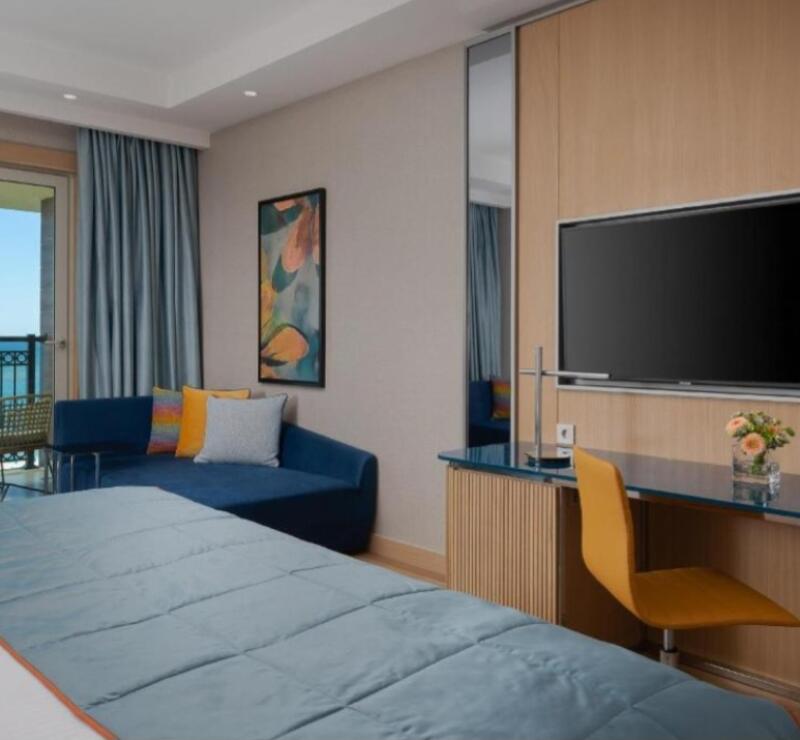 A hotel room features a blue bedspread, a blue sofa with colorful pillows, a desk with a yellow chair, and a TV mounted on the wall.