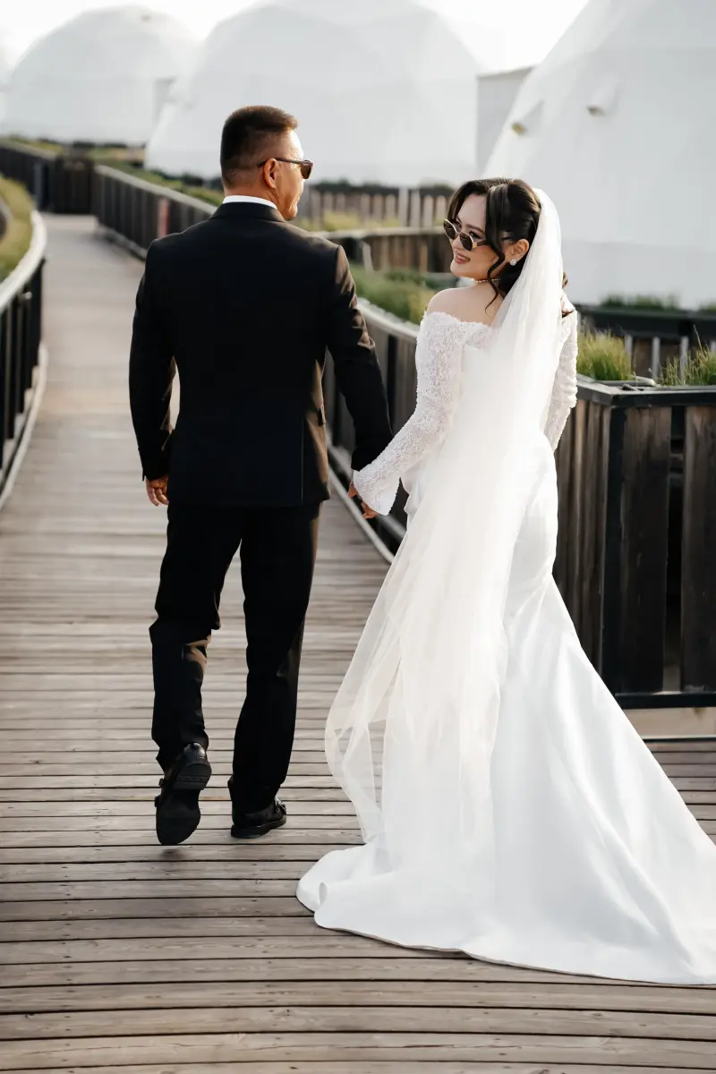 A couple, dressed for a wedding, walks hand-in-hand on a wooden walkway, with white dome structures in the background. The bride, wearing sunglasses and a veil, looks back and smiles.