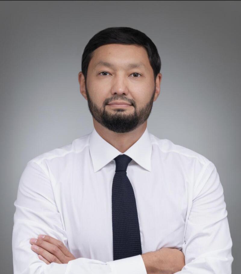 A man with a beard is wearing a white shirt and a dark tie, standing with his arms crossed against a gray background.