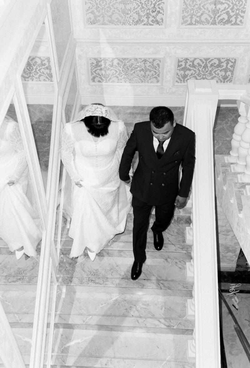 In a black and white photo, a bride in a long white dress and veil walks down marble stairs with a groom in a dark suit.