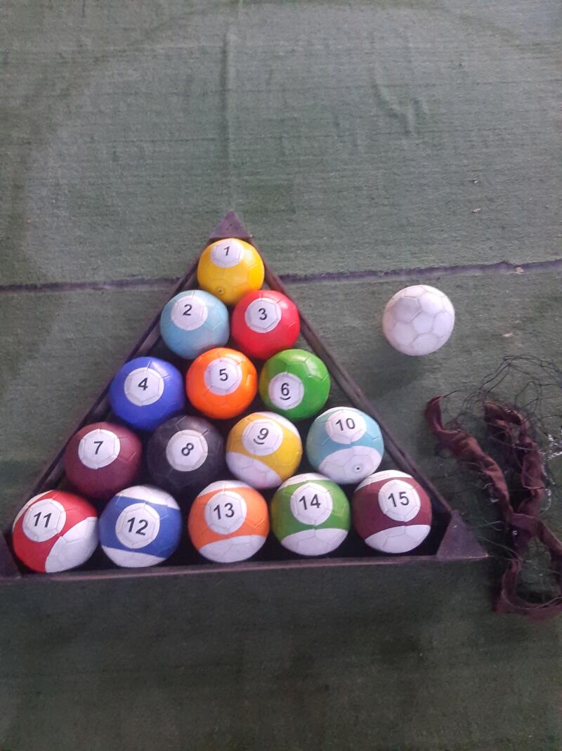 A set of soccer-ball-patterned billiard balls numbered 1 through 15 are arranged in a triangle rack, with a white cue ball lying outside the rack on a green surface.