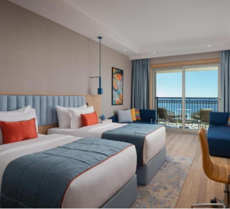 A hotel room features two twin beds with blue and orange accents, a seating area with a blue couch, and a balcony overlooking the ocean.