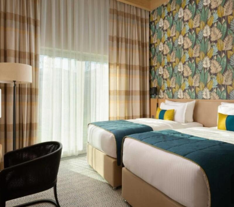 A hotel room features two twin beds with blue and yellow accents, set against a wall with a leafy wallpaper design, complemented by striped curtains and a black chair.