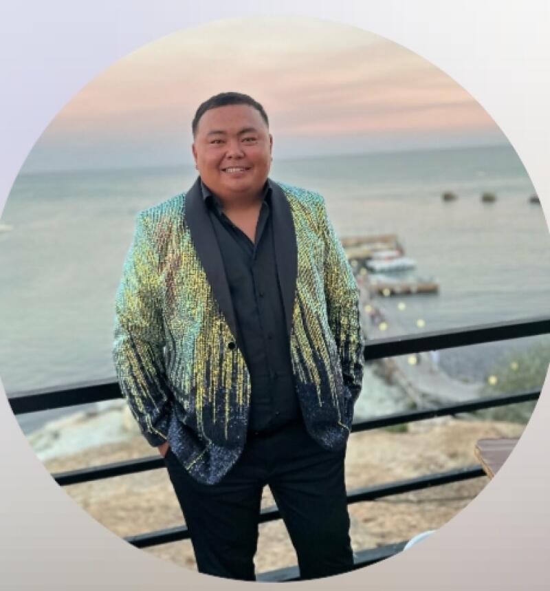 A man in a shiny blazer and black shirt and pants poses in front of a body of water.