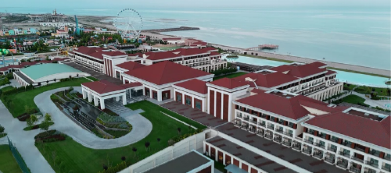 Aerial view of a large resort complex with red roofs, green lawns, and a water feature in the foreground; a Ferris wheel and beach are visible in the background.