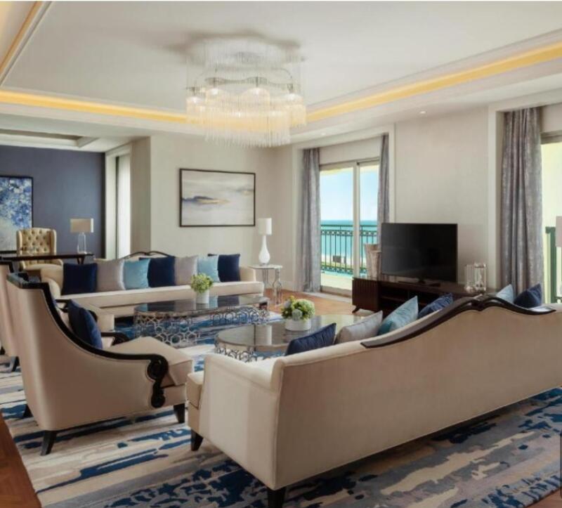 The living room features sofas and armchairs in neutral tones with blue cushions, a glass coffee table, and a patterned blue and white rug. A large chandelier hangs from the ceiling, and sliding glass doors offer a view of the ocean.