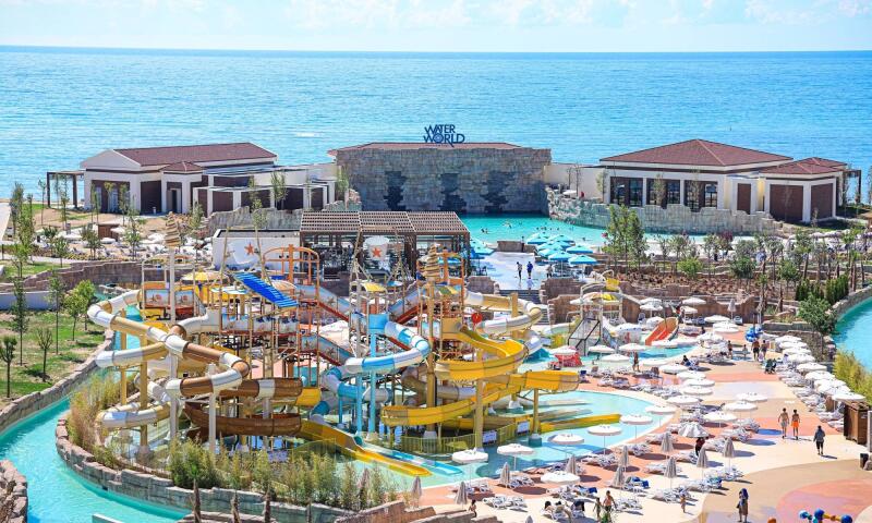 The image shows a large water park complex with colorful slides, pools, umbrellas, and lounge chairs, set against a backdrop of the ocean and buildings, with the words "WATER WORLD" visible in the background.