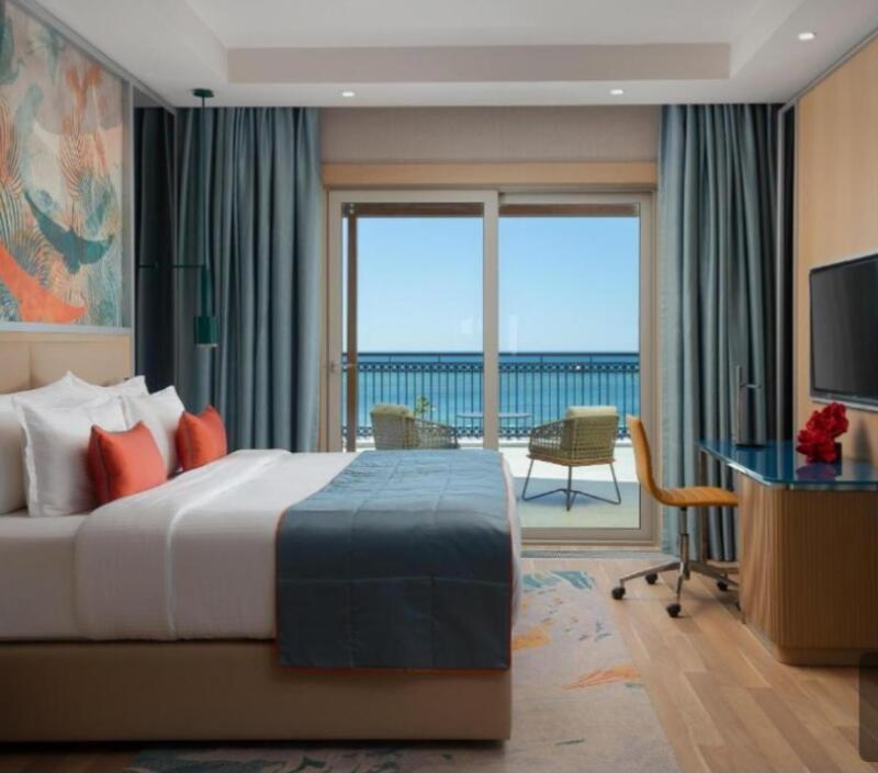 The interior of a hotel room features a bed with white linens and orange pillows, a desk with a chair, and a balcony with a view of the ocean.