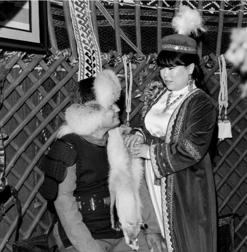 A grayscale image shows a couple in traditional Kazakh clothing holding hands; the man is wearing a fur hat and a vest, while the woman is wearing a decorated robe and a hat with a feather.