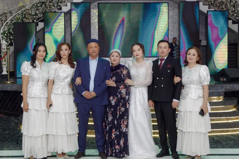 A group of seven people are posing for a photo in formal attire, likely at a wedding or formal event, with a backdrop featuring abstract designs and lighting.
