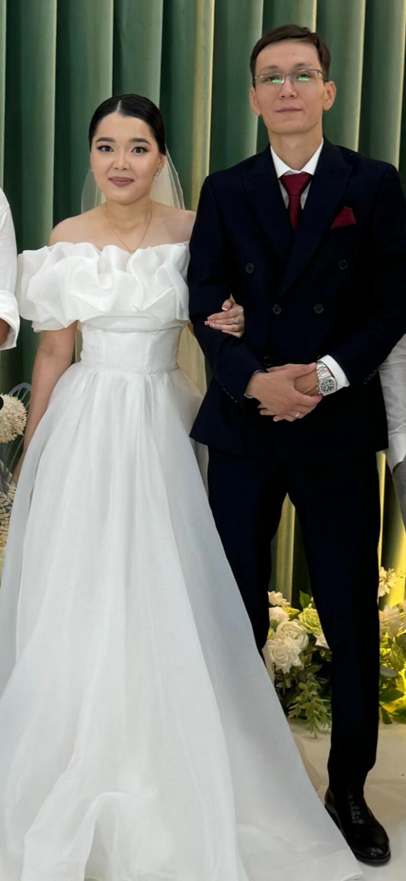 A bride in a white, off-the-shoulder gown stands next to a groom in a dark suit with a red tie and pocket square.