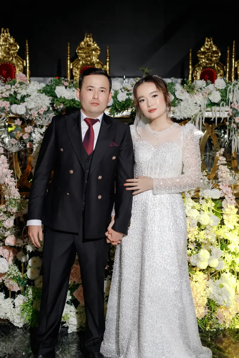 A wedding photo shows a bride in a white gown and a groom in a black suit holding hands in front of a floral backdrop with two gold chairs.