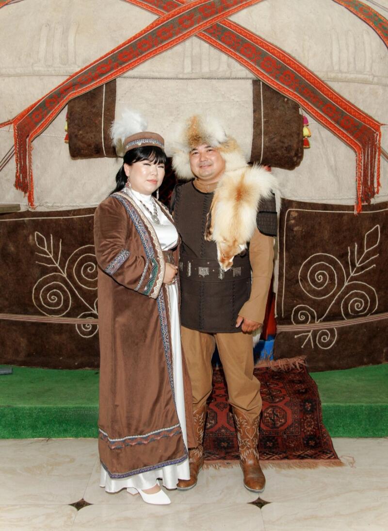 A man and woman are standing in front of a yurt, dressed in traditional Kazakh clothing, with the man wearing a fur hat and vest, and the woman wearing a long brown robe and headdress.