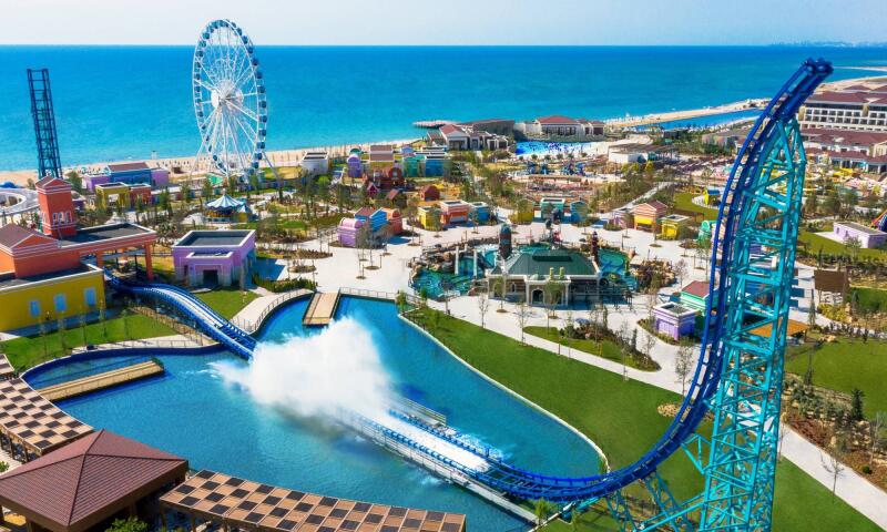 The image shows an amusement park by the sea, featuring a Ferris wheel, colorful buildings, and a water ride with a splash.