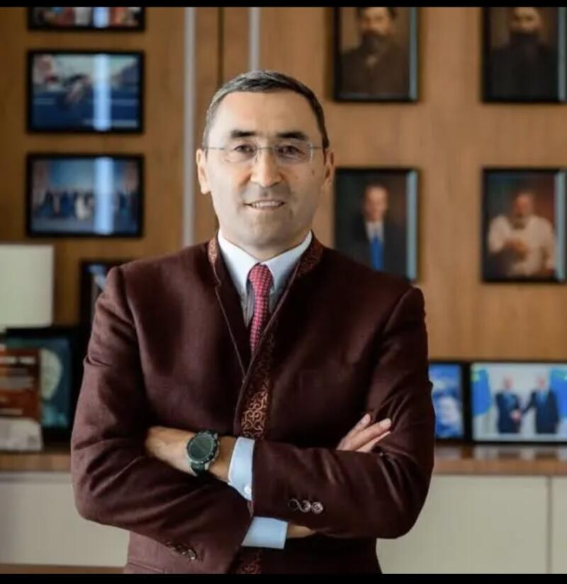 A man in a burgundy suit and glasses stands with his arms crossed, with framed pictures visible in the background.