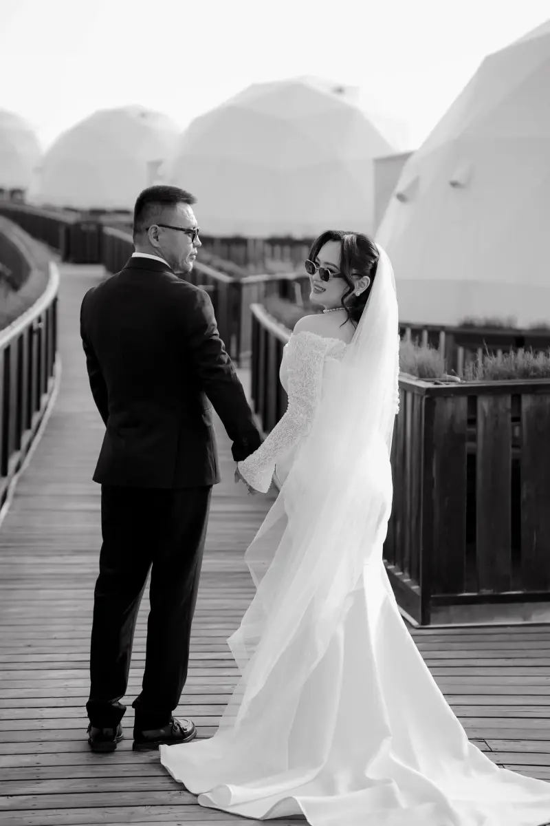 A black and white photo shows a groom in a suit and a bride in a white wedding dress, holding hands and walking on a wooden boardwalk. The bride, wearing sunglasses and a veil, smiles back at the camera while the groom looks towards her, with white dome-shaped buildings in the background.