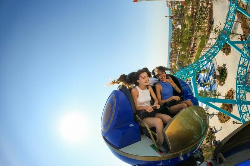 Two women are riding a blue roller coaster under a clear sky, with a view of the park and ocean in the background.