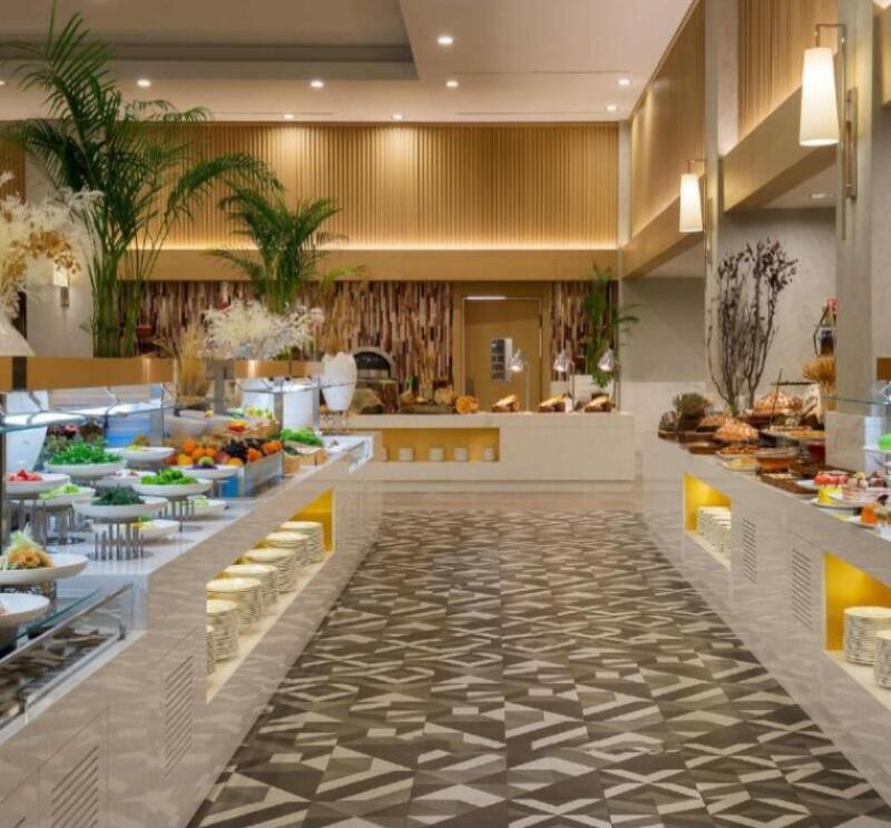 A buffet line with various food options is displayed in a brightly lit room with a patterned floor.