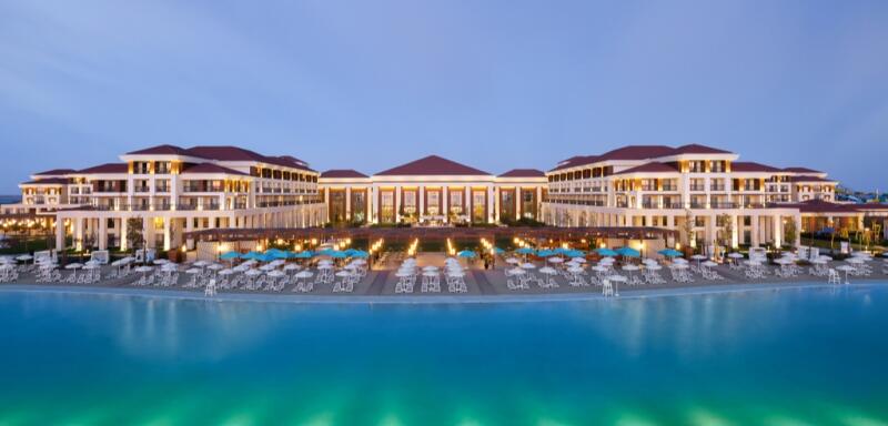 A large resort complex is seen at dusk, featuring multiple buildings with red roofs, white walls, and illuminated windows, positioned behind rows of beach umbrellas and chairs facing a large, turquoise pool.