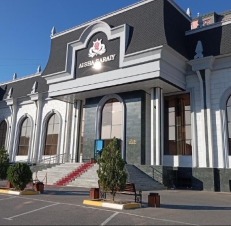 A full shot of Aisha Saraiy, a light-colored building with a dark roof, featuring arched windows and a red carpet leading up to the entrance, under a clear sky.
