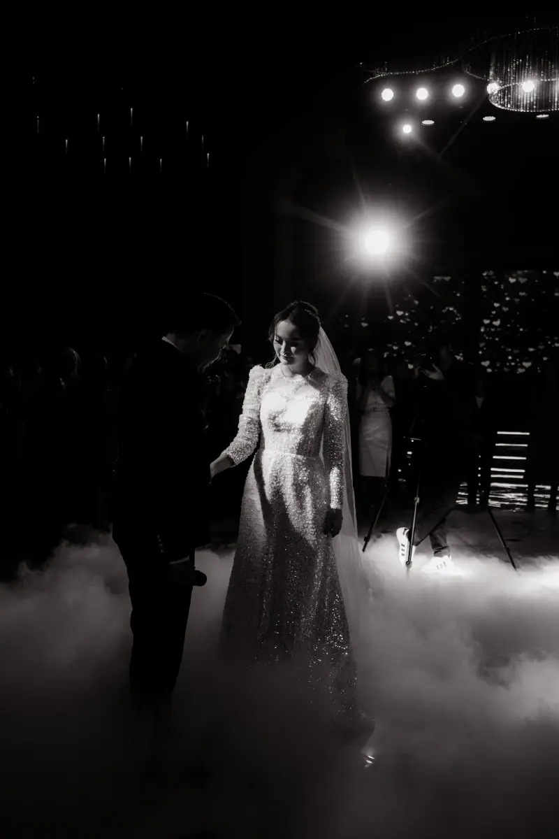 In a black and white photo, a bride in a sparkling gown and veil stands with a groom amidst fog, under bright lights.