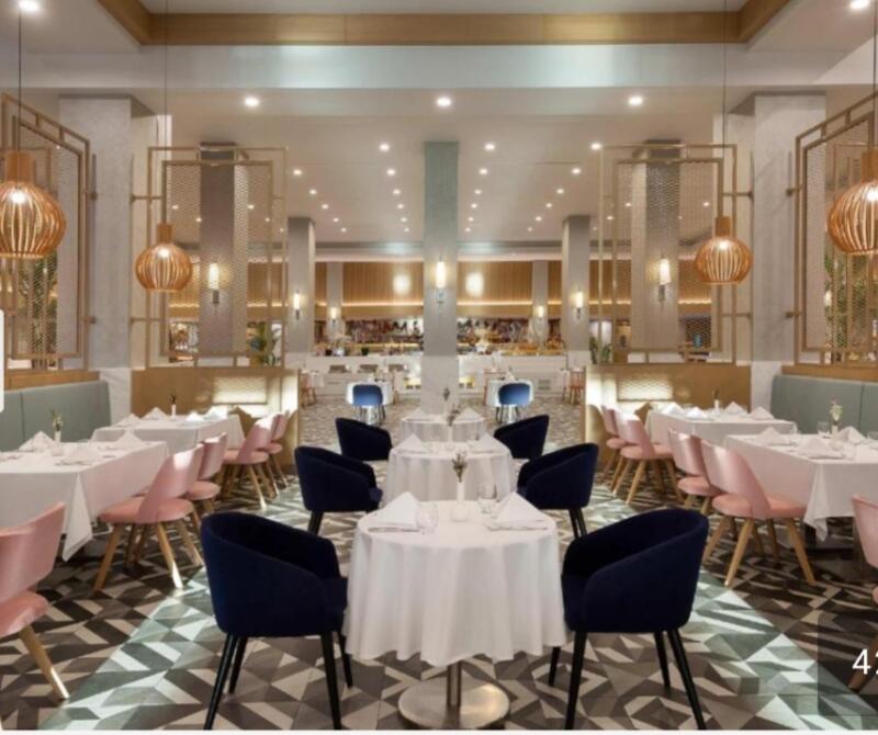 The photo shows a dining room with tables set for a meal, featuring pink and blue chairs, white tablecloths, and patterned flooring.