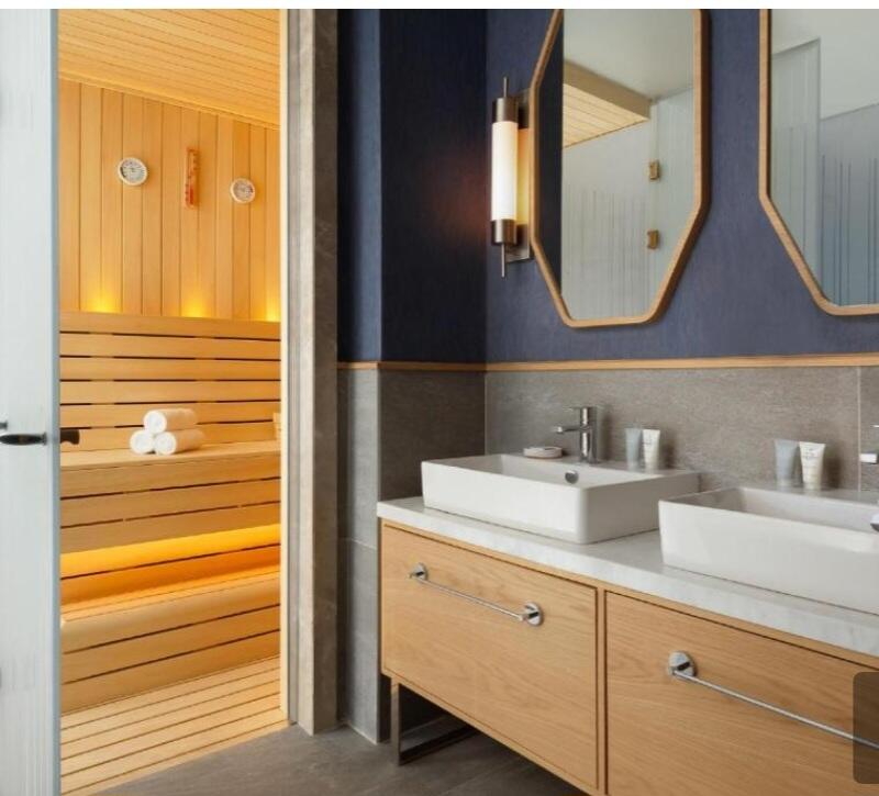 The image shows a bathroom with a sauna, featuring a double sink vanity with a white countertop and wooden cabinets. The sauna is visible through an open doorway, highlighting wooden benches, and towels inside.