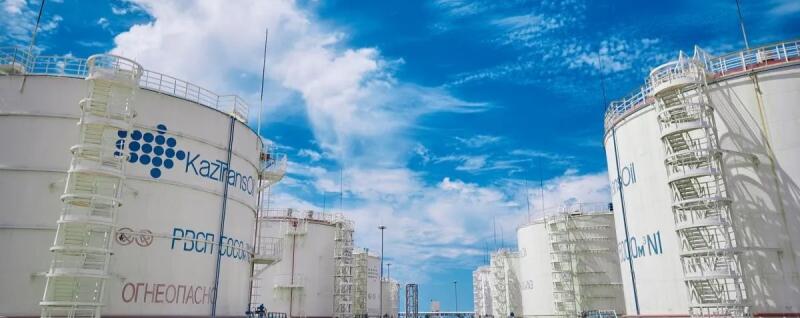 Several large white industrial storage tanks, one prominently featuring the blue 'KazTransOil' logo and the Russian text 'ОГНЕОПАСНО' (FLAMMABLE), stand under a bright blue sky with white clouds.