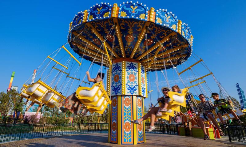 The image shows a swing carousel in an amusement park with people riding on it under a bright blue sky.
