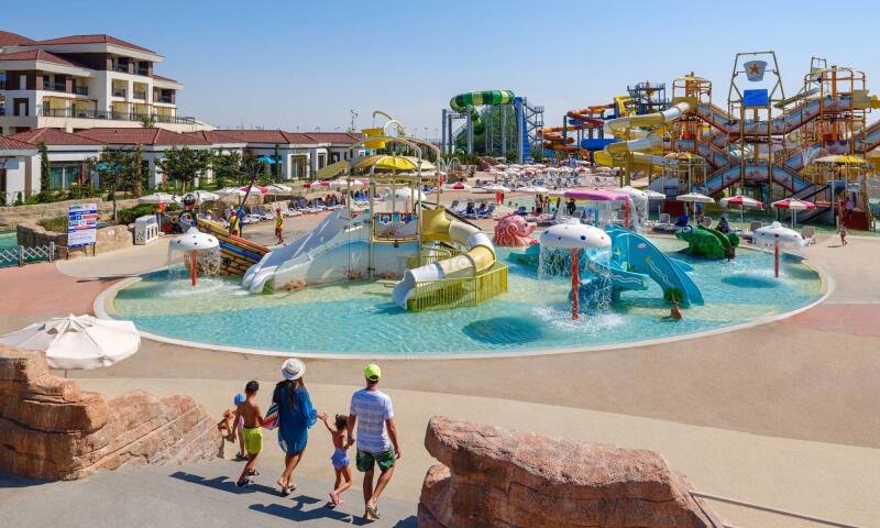 A family walks towards a large water park with various slides and water features under a clear sky.
