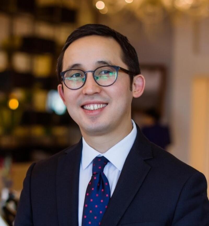 A smiling man with glasses and a dark suit and tie is photographed indoors.
