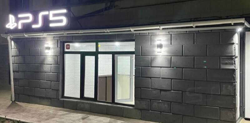 A small store with the PlayStation 5 logo above the door features a gray brick facade and a large window.