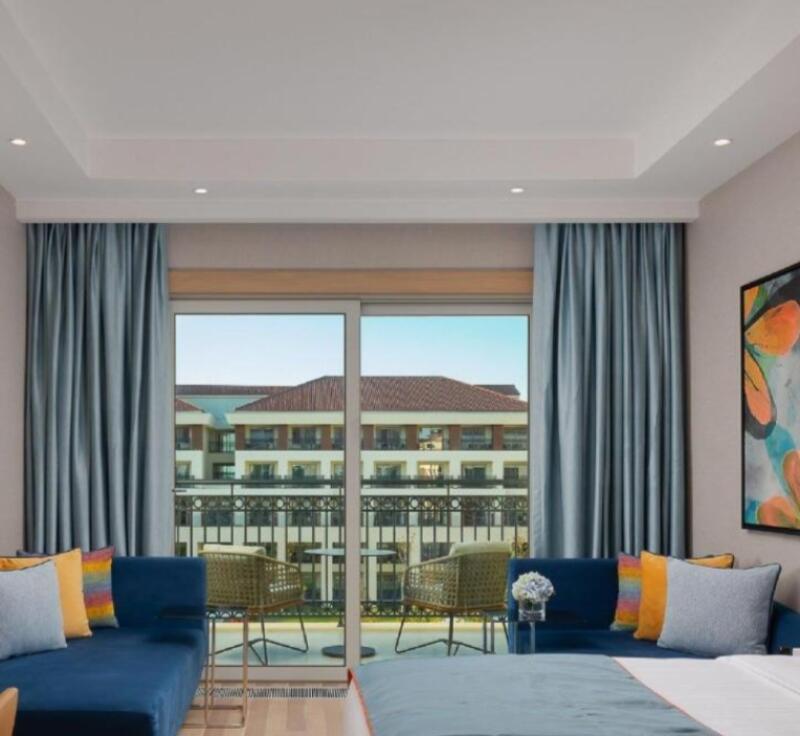 A hotel room features blue sofas with colorful pillows, two wicker chairs on a balcony overlooking a building, and blue curtains.