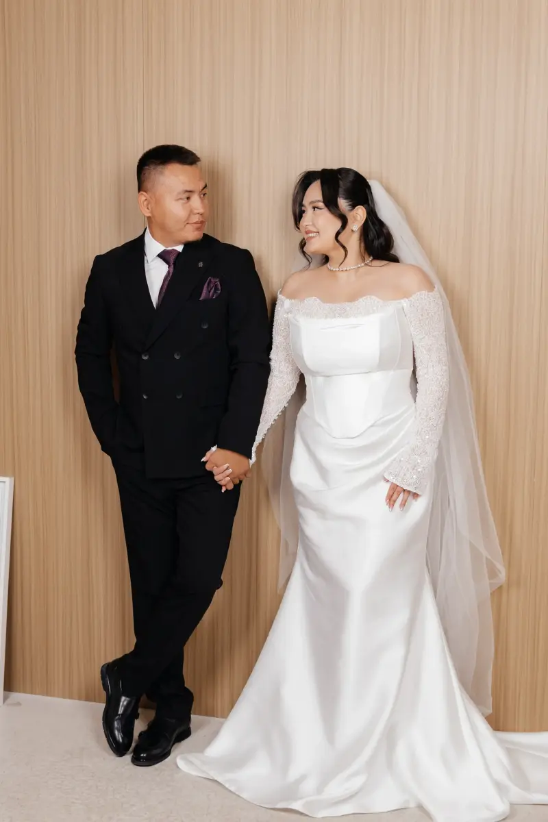 A wedding couple stands, holding hands and looking at each other, against a light brown paneled wall. The groom wears a black suit and the bride wears a white off-the-shoulder gown with a veil.
