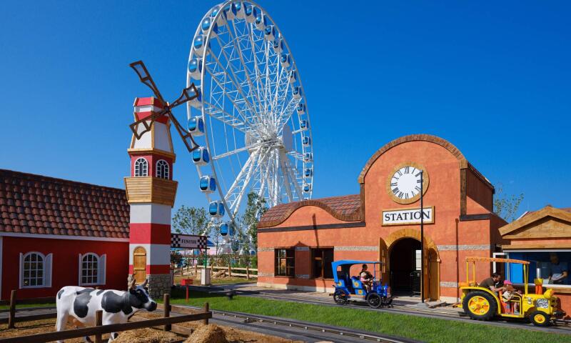 The image shows a park with various attractions, including a Ferris wheel, a windmill, a cow statue, and a train station with a clock and the word "STATION" on it.
