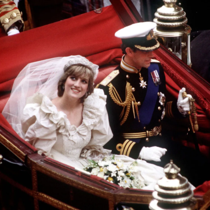 Princess Diana, in a white wedding dress and veil, is sitting next to Prince Charles, who is in a dark military uniform with a white hat, inside of a red carriage.