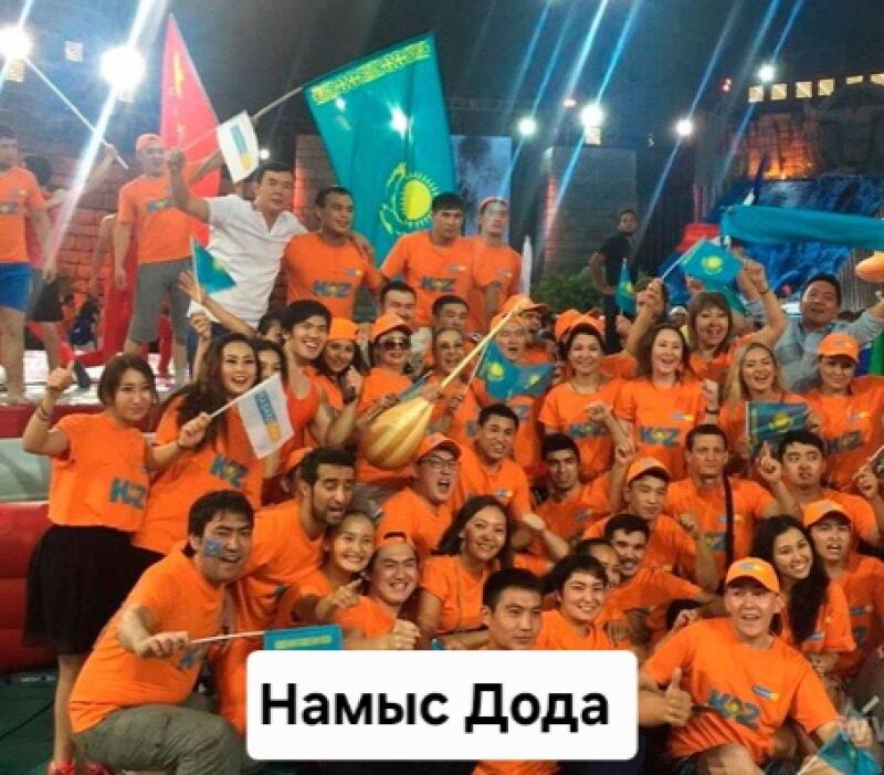 A group of people wearing orange shirts with "KZ" on them are holding Kazakhstan flags and standing together; the text "Намыс Дода" is in the foreground.