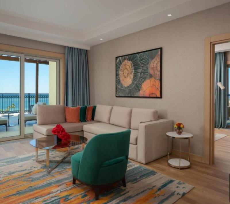 A living room features a beige L-shaped sofa with colorful pillows, a round glass coffee table with red flowers, and a teal accent chair, with a view of the ocean from the balcony.