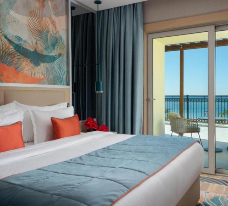 A bedroom features a bed with white linens, orange pillows, and a blue comforter, with a balcony overlooking the ocean.