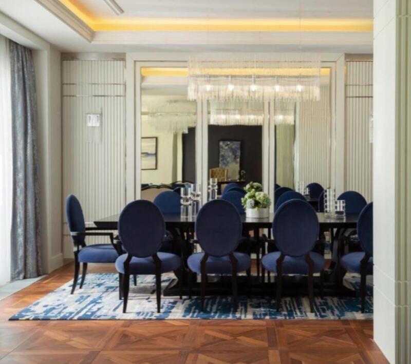 The image shows a dining room with a long, dark table surrounded by blue chairs, set on a blue and white patterned rug over a parquet floor. A chandelier hangs above the table, and the room is decorated with mirrors and neutral-toned walls.