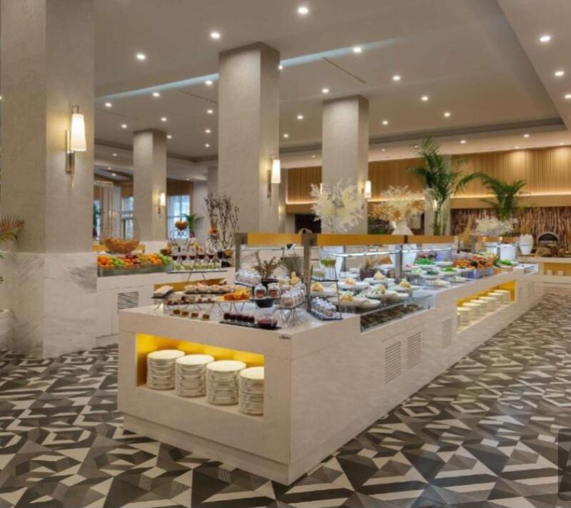 The image shows a buffet with various food items, including fruits, desserts, and salads, displayed on a long, illuminated counter in a spacious hall with patterned flooring and large columns.