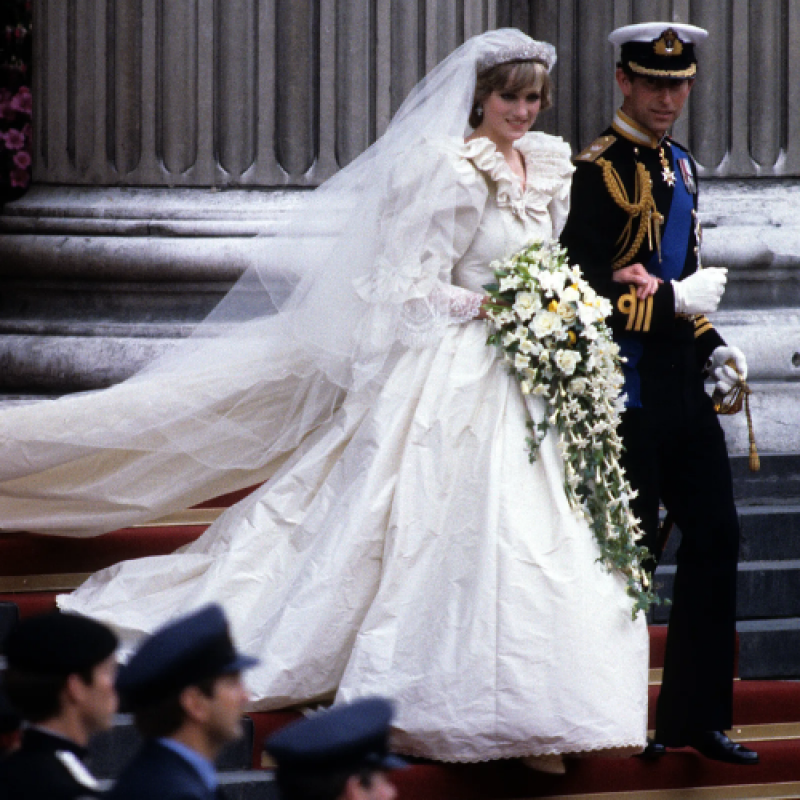 Princess Diana, wearing a white wedding dress with a long train and veil, is walking arm-in-arm with Prince Charles, who is in a navy military uniform with gold accents.