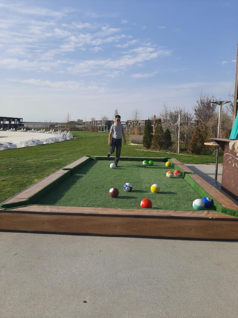 A man in a gray shirt walks toward the camera on a large outdoor pool table with soccer balls instead of billiard balls.